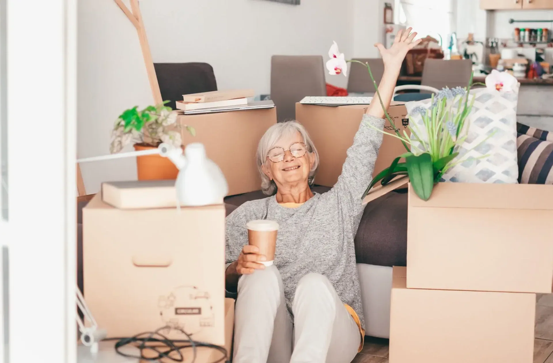 A smiling silver-haired woman in glasses sitting among moving boxes, coffee in hand, one arm raised
