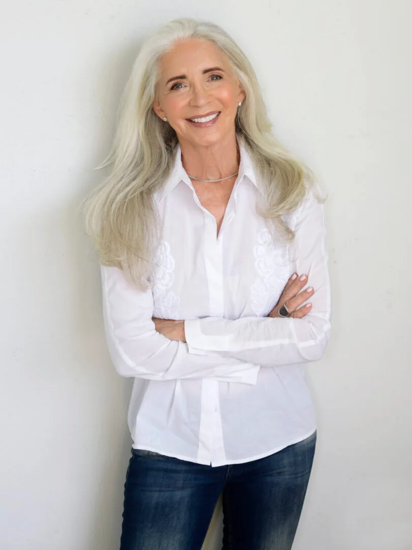 A woman with long silver hair in a crisp white shirt and jeans, arms folded, smiling