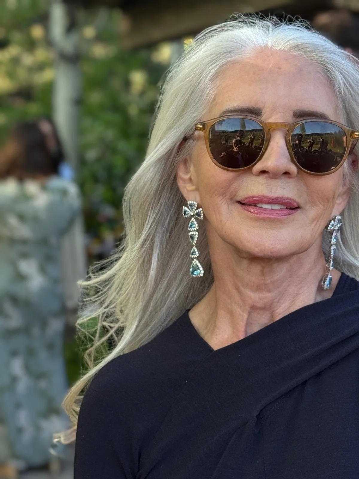 A silver-haired woman in amber sunglasses, aquamarine drop earrings and a black wrap top, outdoors