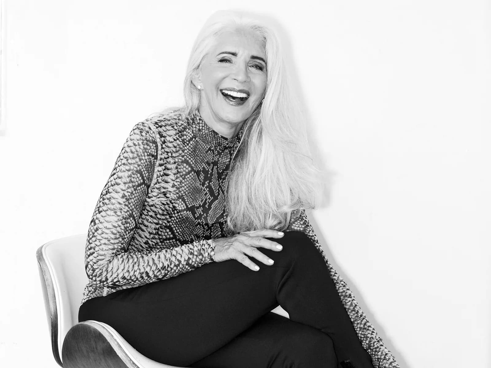 Ruth laughing in a snake print top and black jeans, sitting on a white chair, black and white